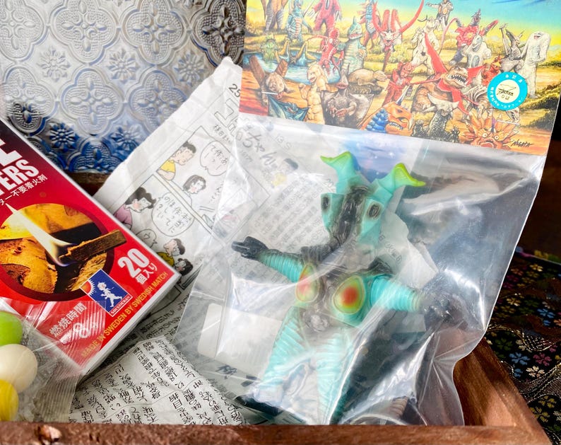 May include: A teal and black monster action figure in a plastic bag, with red and yellow accents. A box of matches with Japanese text and a comic strip are also visible.