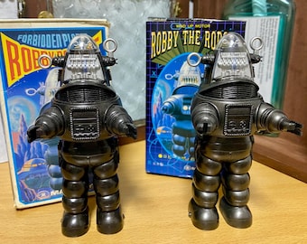 Peut inclure: Deux figurines identiques de Robby the Robot à remonter, de couleur noire, avec des têtes en dôme transparent, des détails internes argentés et des antennes en forme d'anneau, devant leurs boîtes en carton marquées Forbidden Planet et Robby the Robot.