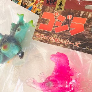 May include: A collection of Godzilla-themed toys, including a translucent pink Godzilla figure and a teal and black monster figure. Packaging includes Japanese text and the CCP 2023 logo.
