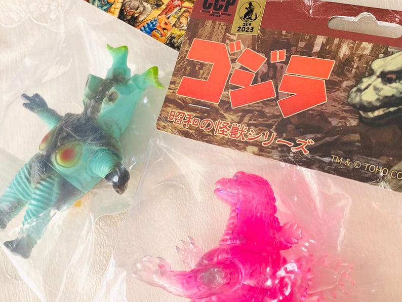 May include: A collection of Godzilla-themed toys, including a translucent pink Godzilla figure and a teal and black monster figure. Packaging includes Japanese text and the CCP 2023 logo.
