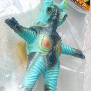 May include: A turquoise and black monster figure with a textured, ribbed design. The figure has a head with horns and outstretched arms. The chest features red and yellow accents. The toy is in a clear plastic bag.