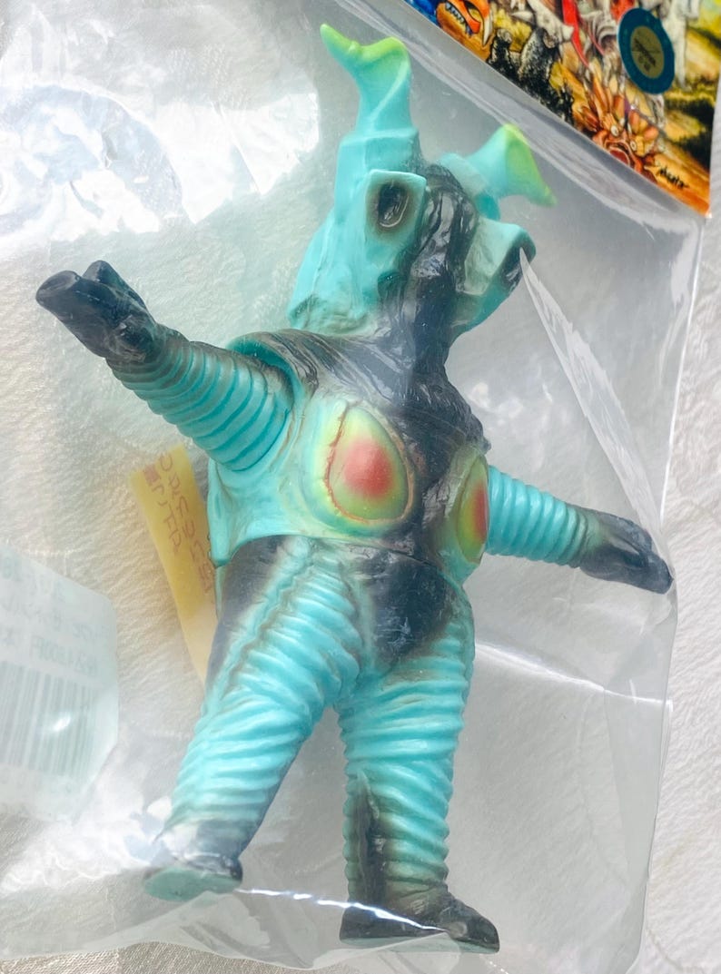 May include: A turquoise and black monster figure with a textured, ribbed design. The figure has a head with horns and outstretched arms. The chest features red and yellow accents. The toy is in a clear plastic bag.