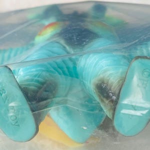 May include: A turquoise Max Toy figure, packaged in clear plastic. The figure has a yellow belly, blue wings, and a rainbow-colored crest. The feet are a lighter shade of turquoise, with the words "Max Toy" printed on one foot.