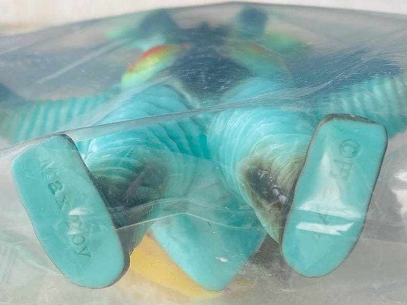 May include: A turquoise Max Toy figure, packaged in clear plastic. The figure has a yellow belly, blue wings, and a rainbow-colored crest. The feet are a lighter shade of turquoise, with the words "Max Toy" printed on one foot.