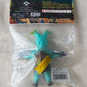 May include: A teal and brown vinyl figure of a monster, packaged in a clear plastic bag. The figure has a unique design with two head protrusions and outstretched arms. The packaging includes Japanese text and a warning label.