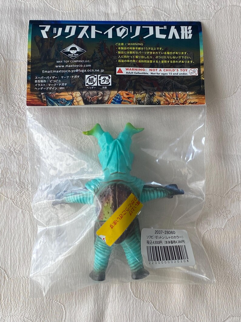 May include: A teal and brown vinyl figure of a monster, packaged in a clear plastic bag. The figure has a unique design with two head protrusions and outstretched arms. The packaging includes Japanese text and a warning label.