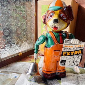 以下が含まれることがあります： オレンジ色のオーバーオールとストライプのシャツを着た、犬の形をしたヴィンテージのぜんまい仕掛けのおもちゃ。 「ANIMAL TIMES」というタイトルの新聞と銀色のベルを持っています。 犬はカラフルな帽子をかぶっています。
