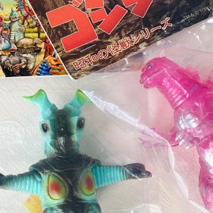 May include: A teal and black monster toy with green horns and a pink Godzilla toy are visible. The toys are in plastic packaging. A colorful illustration of other monsters is in the background.