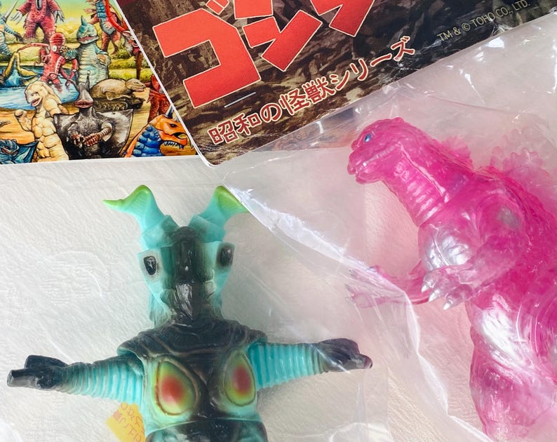 May include: A teal and black monster toy with green horns and a pink Godzilla toy are visible. The toys are in plastic packaging. A colorful illustration of other monsters is in the background.
