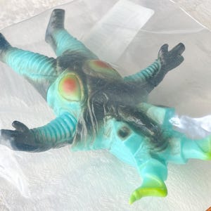 May include: A teal and black monster toy figure with red and yellow accents, packaged in a clear plastic bag. The figure has a detailed design with textured sections and is lying down.
