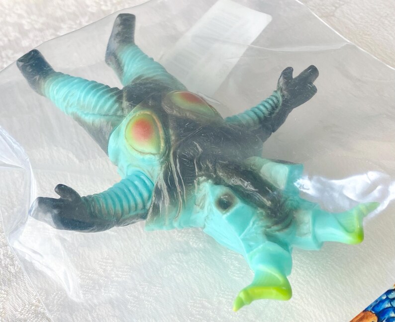 May include: A teal and black monster toy figure with red and yellow accents, packaged in a clear plastic bag. The figure has a detailed design with textured sections and is lying down.