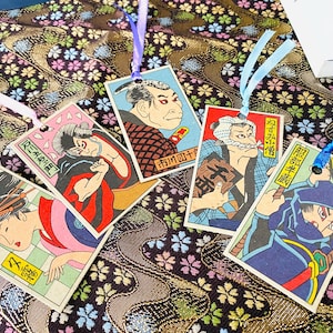 May include: A collection of five Japanese-style bookmarks featuring colorful illustrations of historical figures. Each bookmark has a different design, with vibrant colors and detailed artwork. Each bookmark is attached to a ribbon.