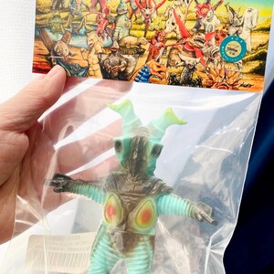 May include: A teal and brown monster figure with a light green head and arms, packaged in a clear plastic bag. The figure has red and orange accents on its chest. The packaging features a colorful illustration of various monsters.