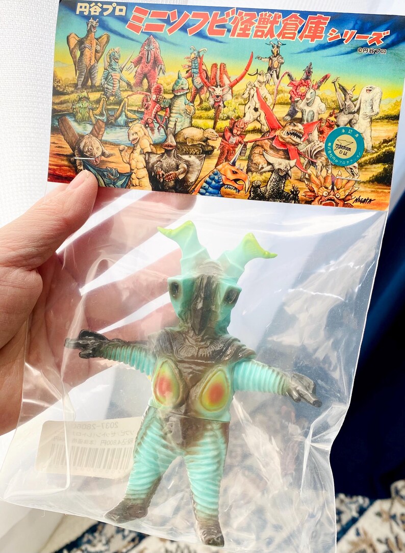 May include: A teal and brown monster figure with a light green head and arms, packaged in a clear plastic bag. The figure has red and orange accents on its chest. The packaging features a colorful illustration of various monsters.