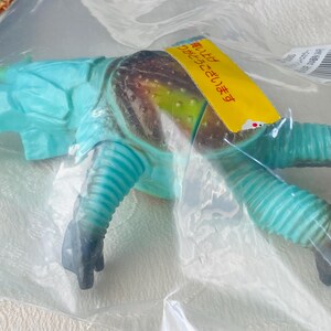 May include: A blue and brown toy creature in a plastic bag. The toy has a segmented body, ribbed legs, and a head with a horn. A yellow tag with Japanese text is attached to the toy.