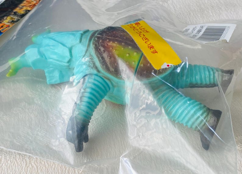 May include: A blue and brown toy creature in a plastic bag. The toy has a segmented body, ribbed legs, and a head with a horn. A yellow tag with Japanese text is attached to the toy.