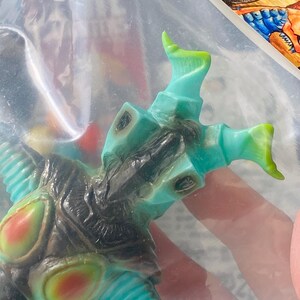 May include: A teal and black monster figurine with green and red accents. The toy is in a clear plastic bag. The figurine has a detailed design with multiple appendages and a textured surface. The overall aesthetic suggests a collectible or novelty item.