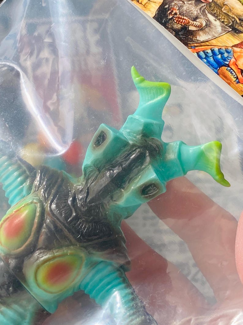 May include: A teal and black monster figurine with green and red accents. The toy is in a clear plastic bag. The figurine has a detailed design with multiple appendages and a textured surface. The overall aesthetic suggests a collectible or novelty item.