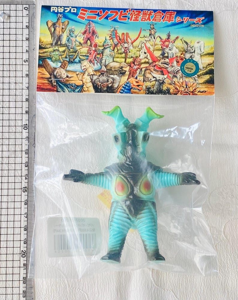May include: A blue and black monster toy figure in a clear plastic bag. The figure has green horns and a light blue body with black accents. The packaging includes Japanese text and illustrations of other monsters.