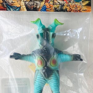 May include: A blue and black monster figure with green horns, packaged in a clear plastic bag. The figure has red and yellow accents on its chest and is part of a series with Japanese text on the packaging.