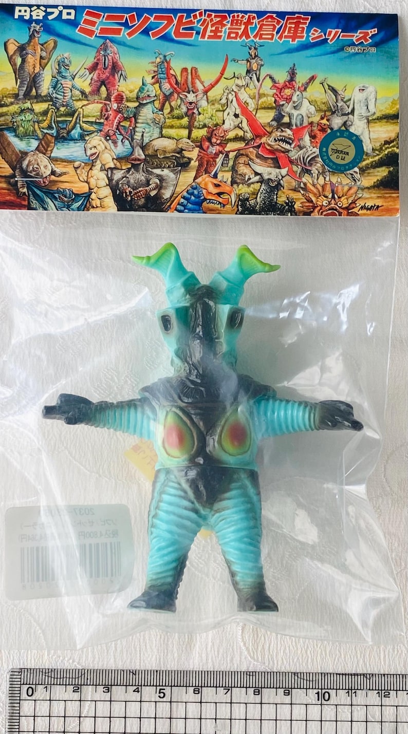 May include: A blue and black monster figure with green horns, packaged in a clear plastic bag. The figure has red and yellow accents on its chest and is part of a series with Japanese text on the packaging.