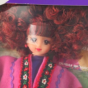 Può includere: Bambola Jenny con capelli ricci rossi e cappotto rosa con finiture floreali. La bambola ha un top viola ed è in una scatola con il testo "Jenny is another yourself" e "Calendar Girl Music".