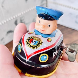 以下が含まれることがあります： 青い帽子をかぶった警官のフィギュアが付いた、ヴィンテージのぜんまい式おもちゃのパトカー。車体は黒と白で、赤いストライプとボンネットには「P.D.」の文字が入っています。おもちゃの側面には巻き上げキーが付いています。