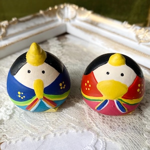 Puede incluir: Dos muñecas japonesas pintadas a mano. Una muñeca es azul con detalles amarillos, la otra es roja con detalles amarillos. Ambas muñecas tienen el pelo negro y están sobre una superficie de encaje blanco.