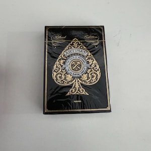 Puede incluir: Una baraja de cartas Artisan, edición negra, con detalles dorados. La parte frontal de la caja presenta un diseño de pica dorado con las palabras "ARTISAN" y "THE GUILD OF ARTISANS" en un emblema circular.