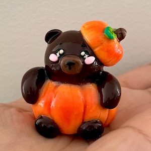 May include: A handcrafted figurine of a brown bear wearing an orange pumpkin hat and costume. The bear has a cute expression with pink cheeks and is holding a pumpkin. This Halloween-themed decoration is perfect for autumn.