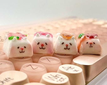 May include: Four custom artisan keycaps in the shape of cute cat faces, each with a different topping. The keycaps are white with pink cheeks and small black facial features. The toppings include strawberries, jelly, and other colorful designs. They are placed on a keyboard.
