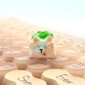 May include: A custom artisan keycap featuring a white dog-like character with a green leafy head covering, pink paws, and a red tongue. The keycap is set on a keyboard with off-white keys, with the "Shift" and "Enter" keys visible.