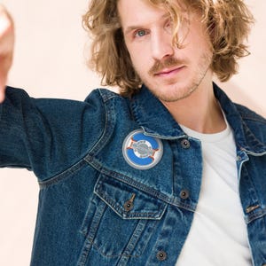 May include: A round, blue and silver pin with a white border is attached to a denim jacket. The pin features a stylized design with a blue outer ring, silver inner details, and a red accent. The jacket is a classic blue denim.