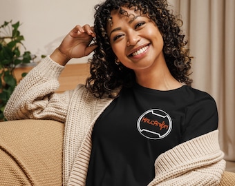 Könnte beinhalten: Eine Frau trägt ein schwarzes T-Shirt mit einem weißen Kreislogo, auf dem "HALCANON" in orangefarbenem Text steht. Sie trägt einen beigefarbenen Cardigan und hat lockiges braunes Haar.