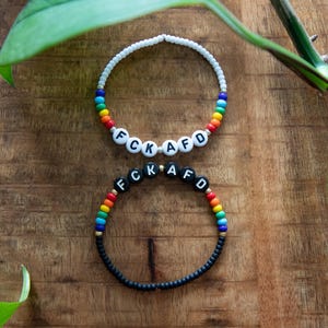 Könnte beinhalten: Zwei Armbänder mit den Buchstaben "FCK AFD". Eines ist weiß mit Regenbogenperlen, das andere schwarz mit Regenbogenperlen. Die Armbänder liegen auf einer Holzoberfläche.