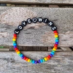 Könnte beinhalten: Ein Armband aus bunten Perlen mit der Aufschrift "LOVE WINS" in schwarzen Buchstabenperlen. Das Armband zeigt einen Regenbogen von Farben, von Rot bis Violett. Ein Herzsymbol ersetzt den Buchstaben "O" in "LOVE".