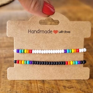 Könnte beinhalten: Zwei Armbänder mit Perlen, eines weiß und eines schwarz, jeweils mit einem Abschnitt in Regenbogenfarben. Die Armbänder werden auf einer braunen Karte mit der Aufschrift "Handmade with love" präsentiert.