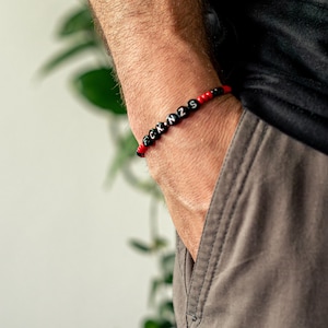 Könnte beinhalten: Ein schwarz-rotes Perlenarmband mit den Buchstaben "FCK N2S" in Weiß. Das Armband wird am Handgelenk getragen, mit einer grauen Tasche im Hintergrund.