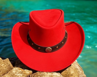 May include: A vibrant red cowboy hat with a brown leather band adorned with rivets and a decorative buckle. The hat is set against a backdrop of turquoise water and rocks, showcasing its bold color and classic design.