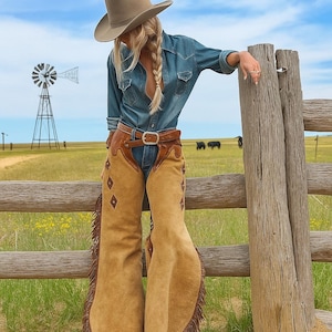 Puede incluir: Camisa vaquera y chaparreras de ante color canela con flecos y diamantes. Un cinturón marrón con hebilla plateada y un sombrero de vaquero color canela completan el look western. Un molino de viento y una valla de madera están en el fondo.