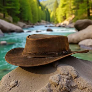 Könnte beinhalten: Ein brauner Lederhut mit breiter Krempe und Band am Kronenansatz. Der Hut liegt auf einer sandigen Oberfläche, mit einem Fluss und Bäumen im Hintergrund. Der Hut scheint neu und ungetragen zu sein.