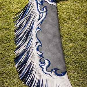 May include: A gray leather chap featuring decorative overlays in white and dark blue. The side panel is adorned with long, layered fringe strips in alternating white and dark blue colors, extending along the entire length of the leg covering.