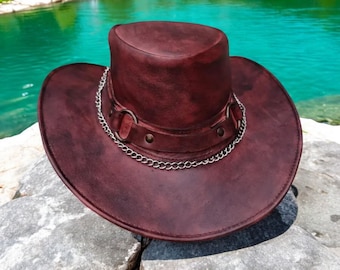 May include: A burgundy leather hat with a wide brim and a cylindrical crown. A silver chain and metal rings encircle the hat. The hat is resting on a gray rock, with a turquoise lake and green trees in the background.