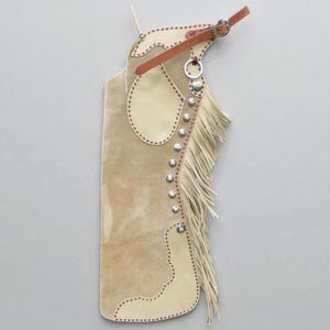 May include: Tan suede leather chaps with cream-coloured accents and fringe. The chaps feature decorative stitching, silver studs, and a brown leather belt with a buckle. A western-style garment.