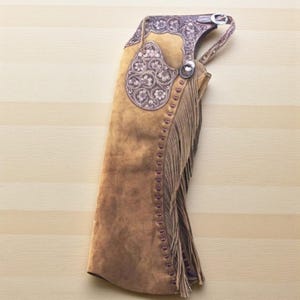 Puede incluir: Chaps de cuero marrón con detalles florales plateados y flecos laterales. Los chaps tienen un cierre de hebilla y están diseñados para montar a caballo. El cuero tiene un aspecto envejecido.
