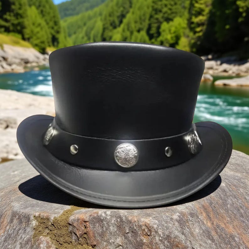 May include: A black leather top hat with a silver band and decorative silver buttons. The hat has a classic top hat shape with a wide brim. The hat is sitting on a rock with a river and trees in the background.