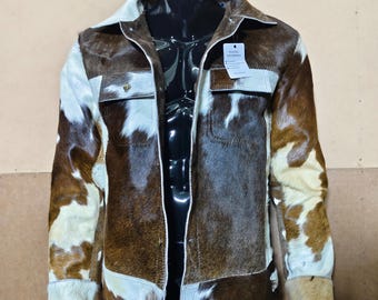 May include: A brown and white cowhide jacket with a collared neckline, two chest pockets, and button closures. The jacket has white accents on the collar, pockets, and cuffs. The jacket is displayed on a black mannequin.