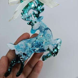 May include: A decorative reindeer ornament with a white ribbon. The reindeer is blue and white with glitter accents. The ornament is approximately 10 cm tall.