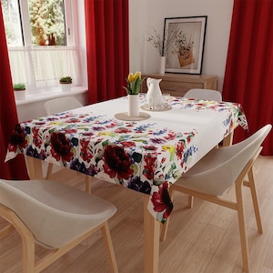Könnte beinhalten: Ein rechteckiges weißes Tischtuch mit einem Blumenmuster aus roten, blauen und gelben Blumen bedeckt einen Holztisch. Der Tisch ist von vier weißen Stühlen umgeben. Rote Vorhänge rahmen ein Fenster im Hintergrund ein.
