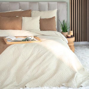 Könnte beinhalten: Ein Bett mit einer cremefarbenen, gesteppten Bettdecke und passenden Kissen. Akzentkissen in Braun- und Cremetönen sind auf dem Bett angeordnet. Ein Holztablett enthält ein Buch, eine Tasse und dekorative Gegenstände. Das Kopfteil ist hellgrau.